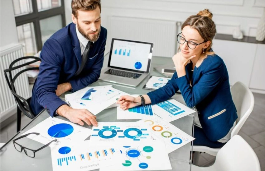 Two professionals discuss marketing data charts and graphs spread on a table with a laptop showing analytics.